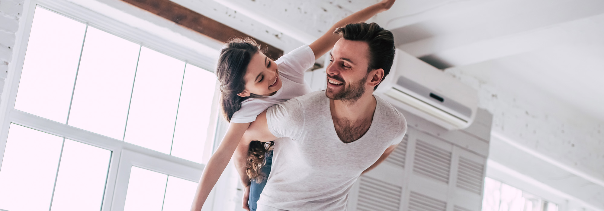 A man playfully gives a piggyback ride to a young girl inside a bright, airy room with large windows. Both are smiling and wearing casual white shirts. The room has white walls and a visible air conditioning unit.