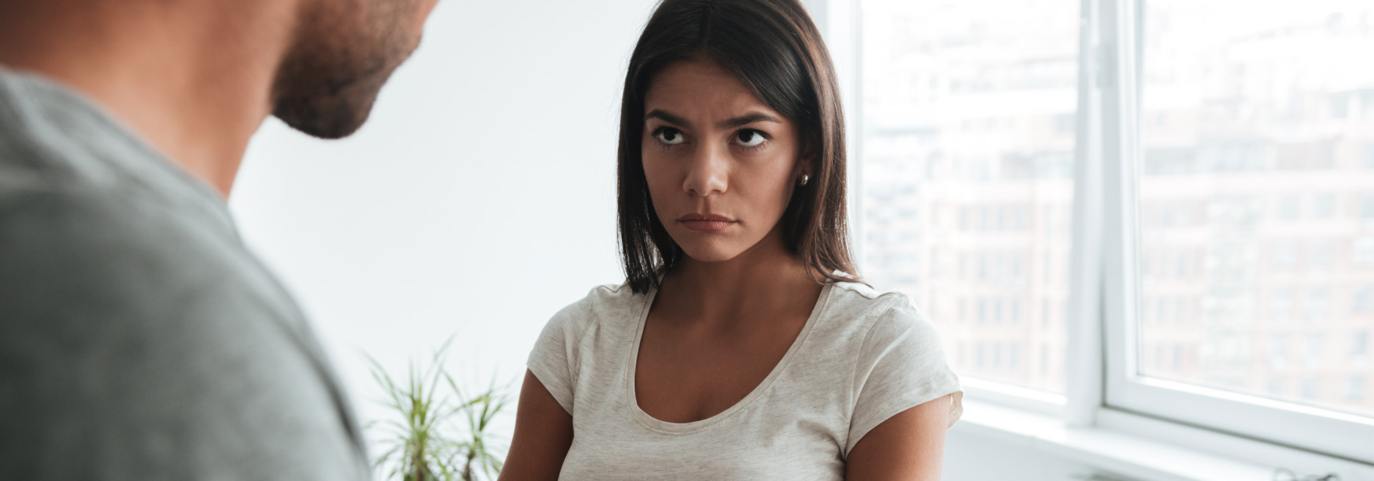 Woman wearing a white shirt stands with arms crossed, looking displeased. She is indoors with a blurred window background and a plant visible.