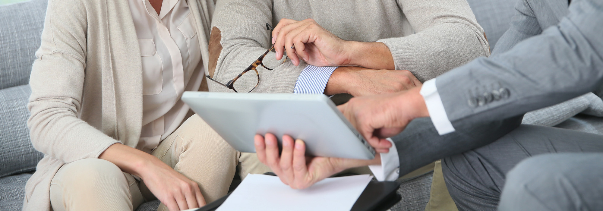 A close-up of three people sitting on a couch, engaged in discussion. One person points to a tablet held by another. Papers and eyeglasses are visible, suggesting a meeting or consultation. They wear business casual attire.
