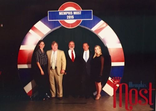Five people standing together and posing in front of a large sign that says "Memphis Most 2015." The background features a design with red, blue, and white sections, resembling a target.
