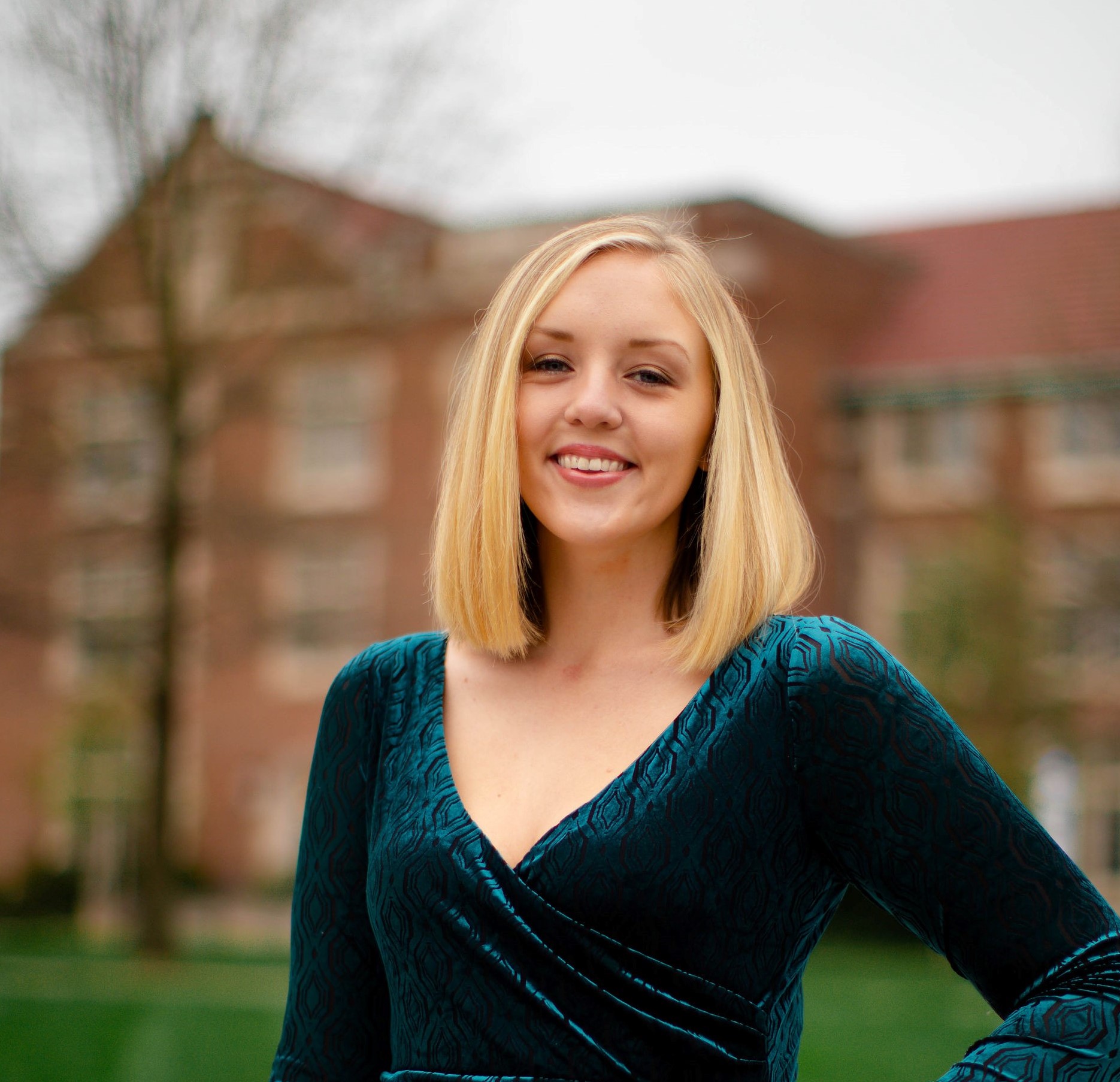 A person with shoulder-length blond hair smiles outdoors, wearing a teal dress. The background is blurred, showing a large brick building and trees.