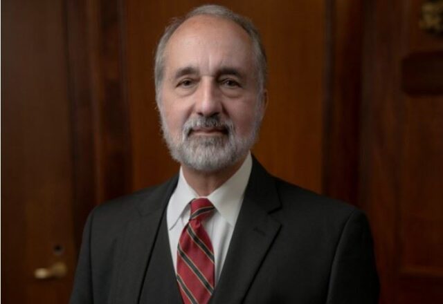 An older man with a beard and mustache is wearing a suit, white shirt, and red striped tie. He stands in front of a wooden background, looking directly at the camera with a neutral expression.