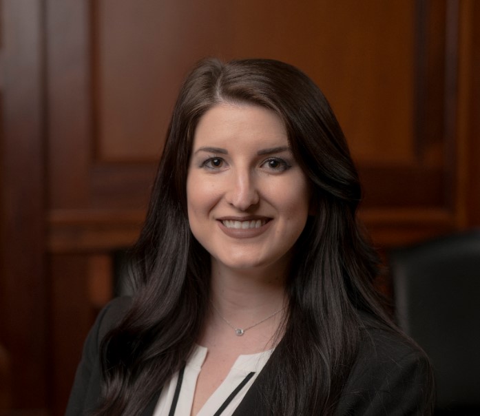 A woman with long dark hair smiles, wearing a black jacket over a white top. She is seated in a room with wooden paneling, giving a professional impression.