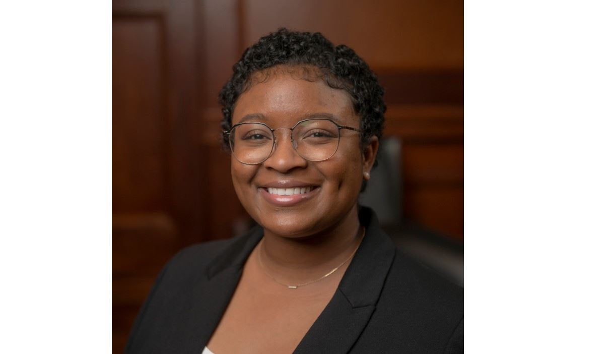 A person with short curly hair and glasses is smiling. They are wearing a black jacket over a white top. The background is a blurred wooden paneled room.