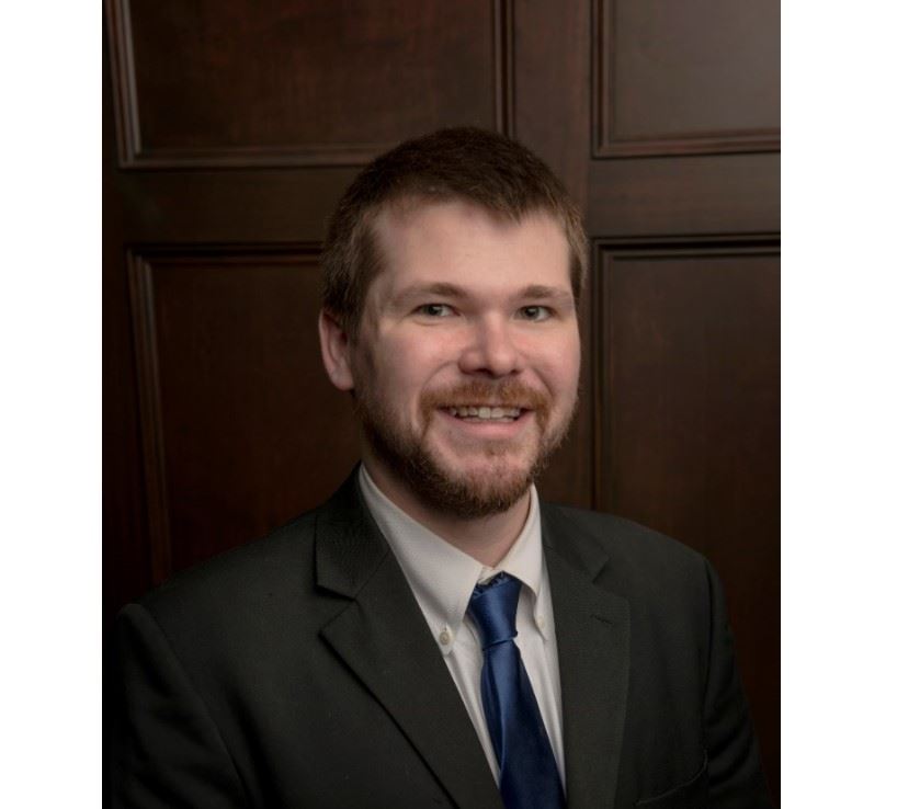 A person with short hair and a beard is smiling while wearing a dark suit, white shirt, and blue tie. They are posed in front of a wooden panel background.