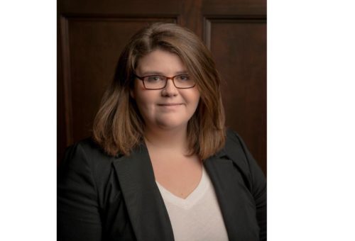 A person with shoulder-length brown hair and glasses is wearing a black blazer over a white shirt, standing against a wooden background.