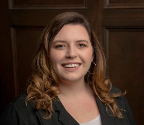 Woman with long, wavy hair and hoop earrings smiling, wearing a dark blazer over a light top. She stands in front of a wooden panel backdrop.