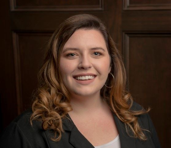 Woman with long, wavy hair and hoop earrings smiling, wearing a dark blazer over a light top. She stands in front of a wooden panel backdrop.