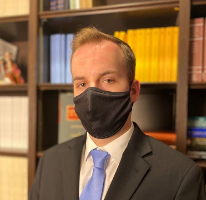 A person wearing a black mask, suit, and blue tie is standing in front of a bookshelf filled with books of various colors.
