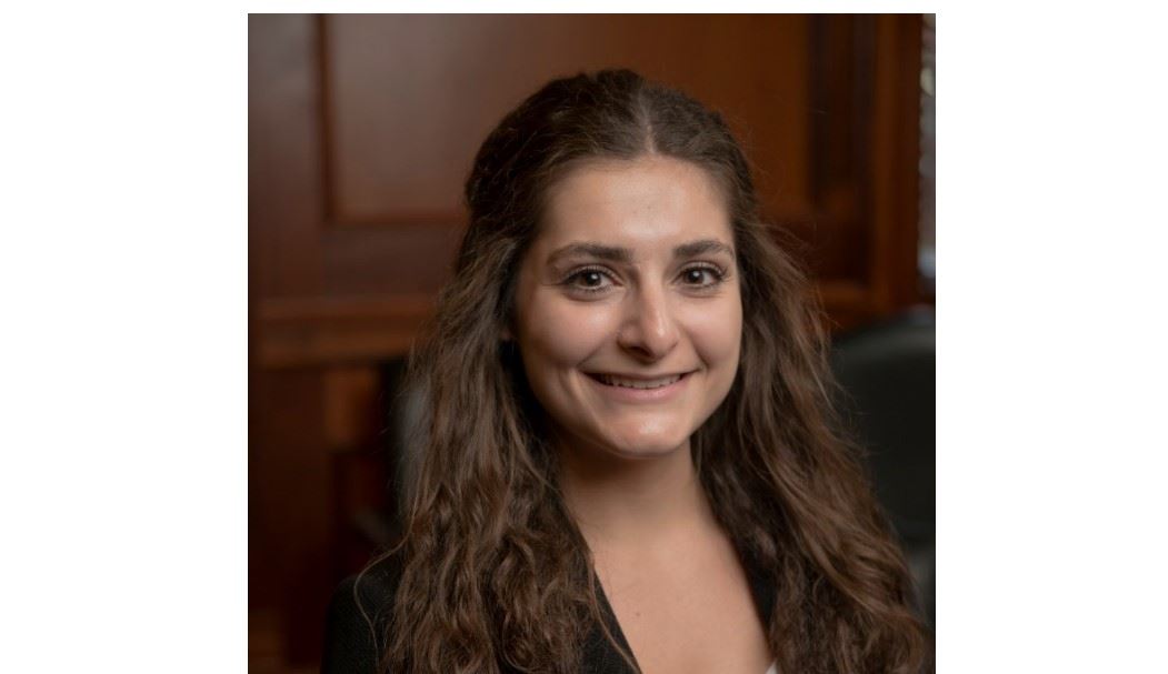 A person with long, wavy brown hair is smiling at the camera. They are wearing a dark blazer over a light top. The background features a wooden paneled wall and a blurred chair.