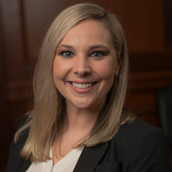 Smiling woman with shoulder-length blonde hair, wearing a white blouse and black blazer, stands against a dark wood background.