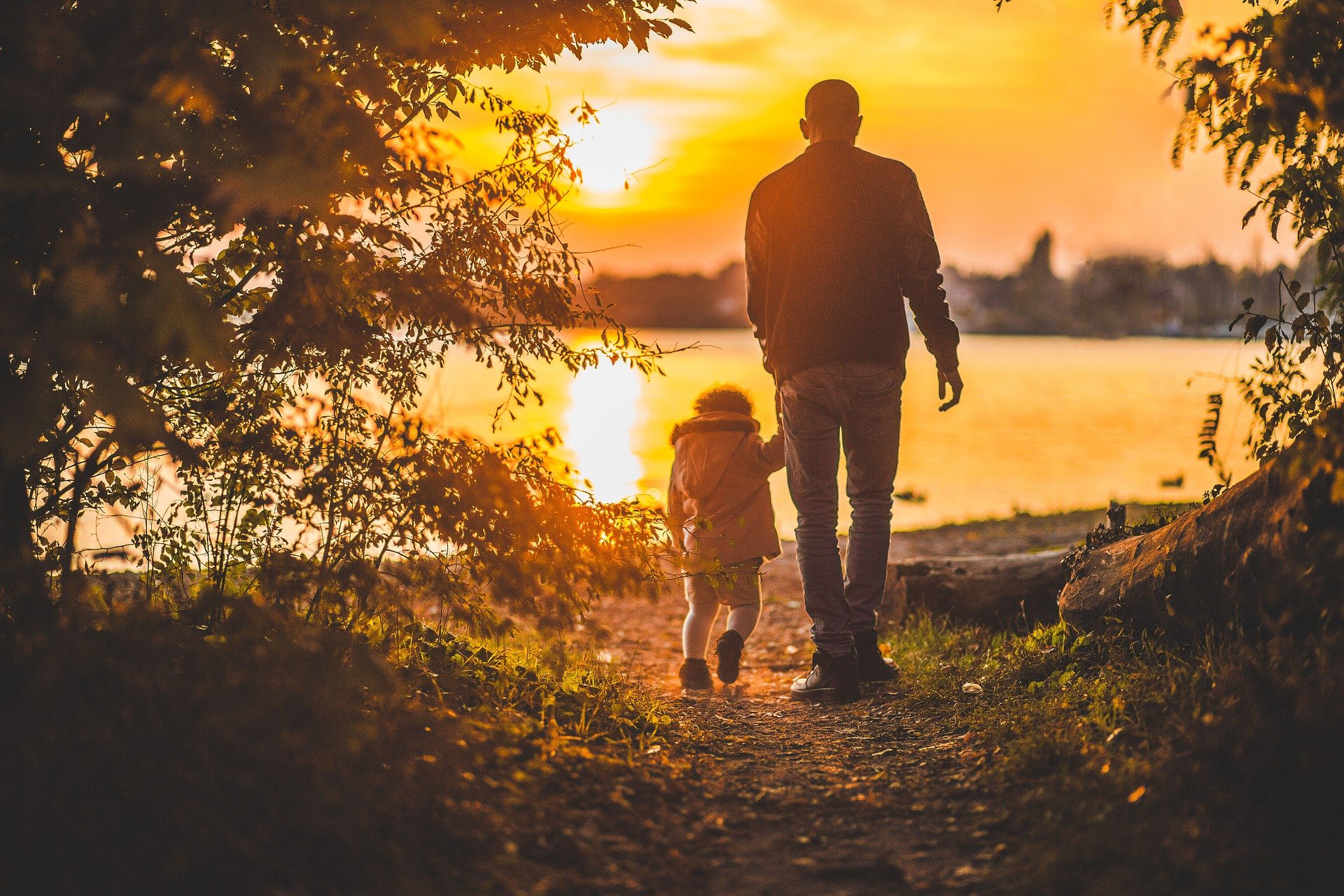 A man and a child walk hand in hand along a path by a lake, framed by trees. The sky is vibrant with warm orange hues from the setting sun, casting reflections on the water and creating a peaceful, picturesque scene—a moment to cherish and protect forever.