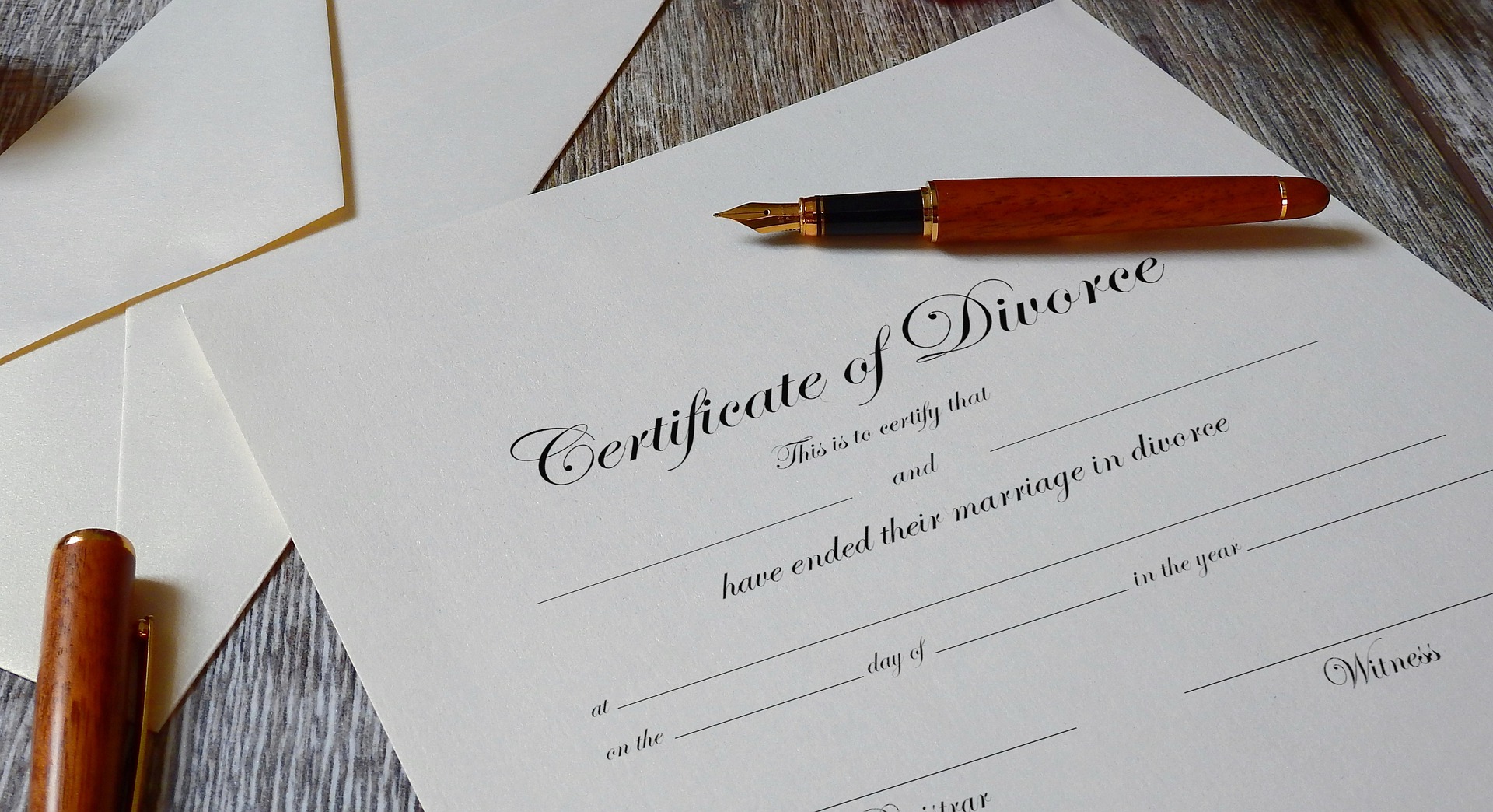 A divorce certificate rests on a wooden table, with a fountain pen poised protectively on top. The paper is partially filled out, leaving spaces for details. Nearby, an envelope awaits its duty.