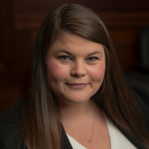 A person with long brown hair wearing a black blazer over a white top is smiling softly. The background is a dark, blurred interior setting.