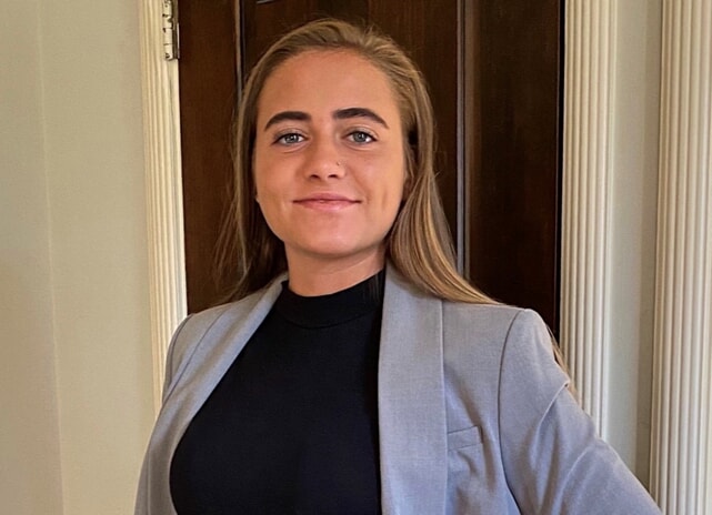 Smiling person with long hair in a gray blazer and black shirt, standing near a wooden doorframe.