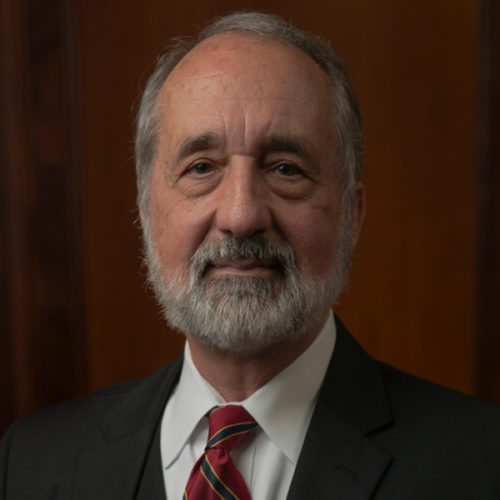 An older man with a beard and mustache is wearing a dark suit, white shirt, and striped tie. The background is a wooden panel.