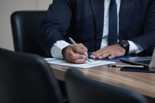 A person in a suit carefully writes on a document at a wooden table, aiming to protect crucial details. Nearby, a laptop and pen await further use in what appears to be an office or conference room.