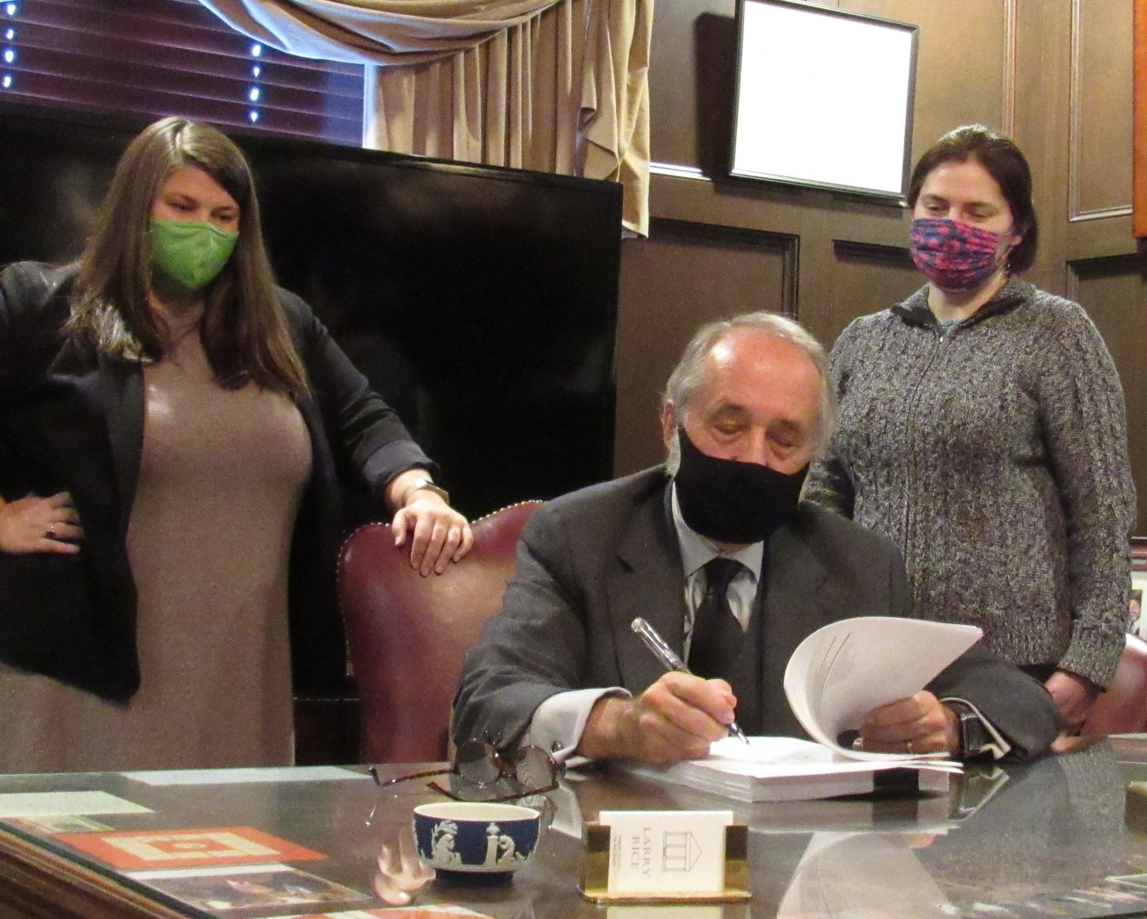 A man in a suit and mask is seated at a desk, signing documents. Two women wearing masks stand beside him, one in a blazer and dress, the other in a sweater. The setting is an office with framed pictures on the walls.