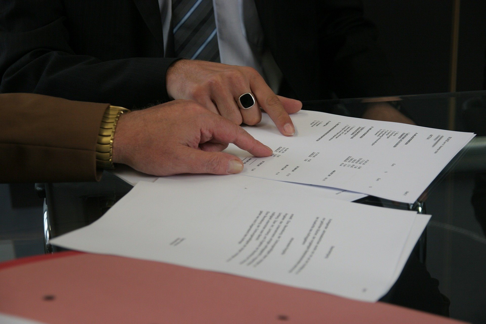 Two people are reviewing credit documents on a glass table. One person points at the papers while the other, dressed in a sleek suit, wears a gold watch and ring.