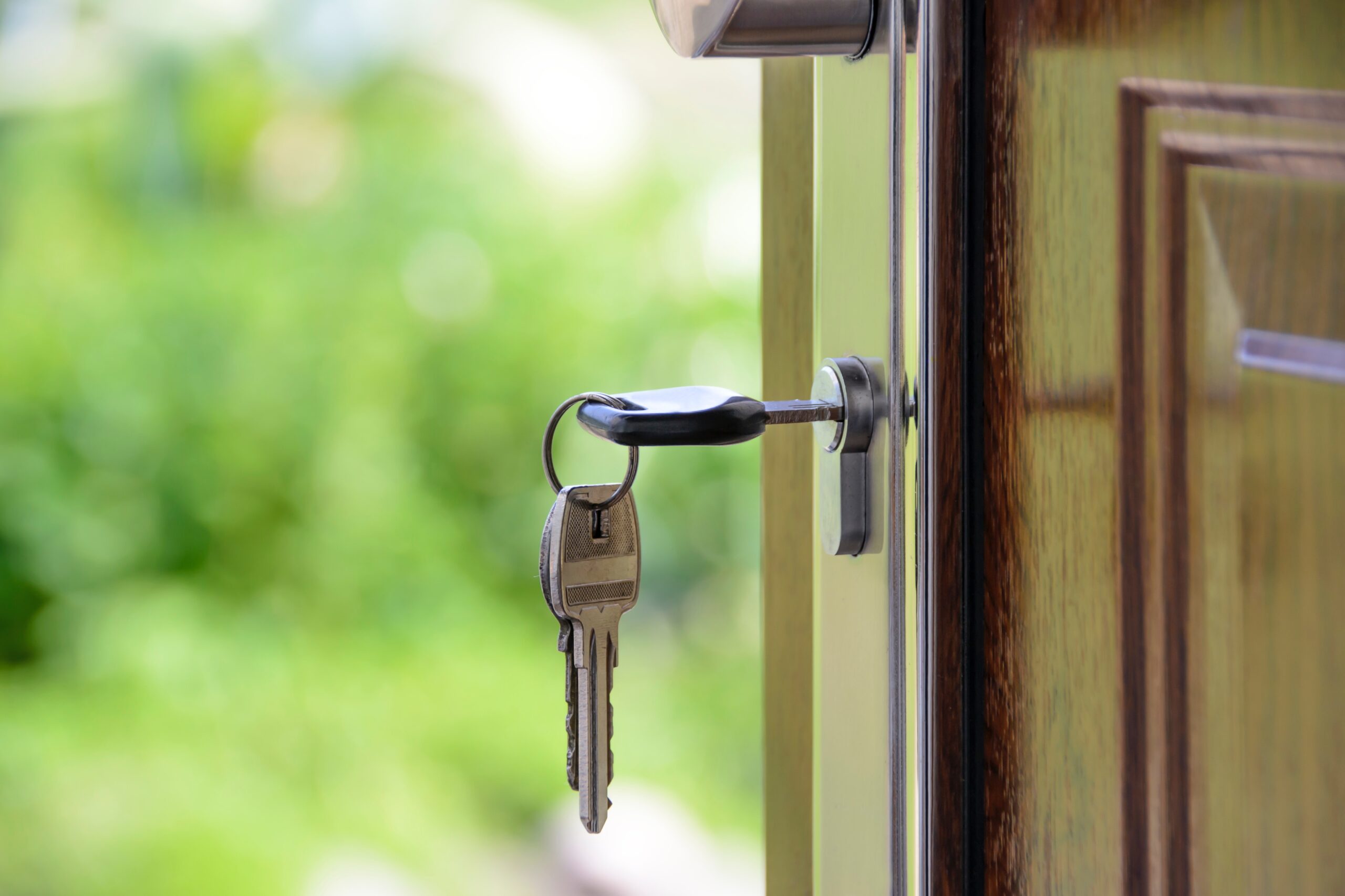 A close-up of a wooden door slightly ajar, with a silver lock sharply in focus, reflects subtle signs of house life and quiet moments post-divorce. Two keys hang from a black keyring, poised in the lock, against a backdrop of blurred green foliage hinting at the garden beyond.