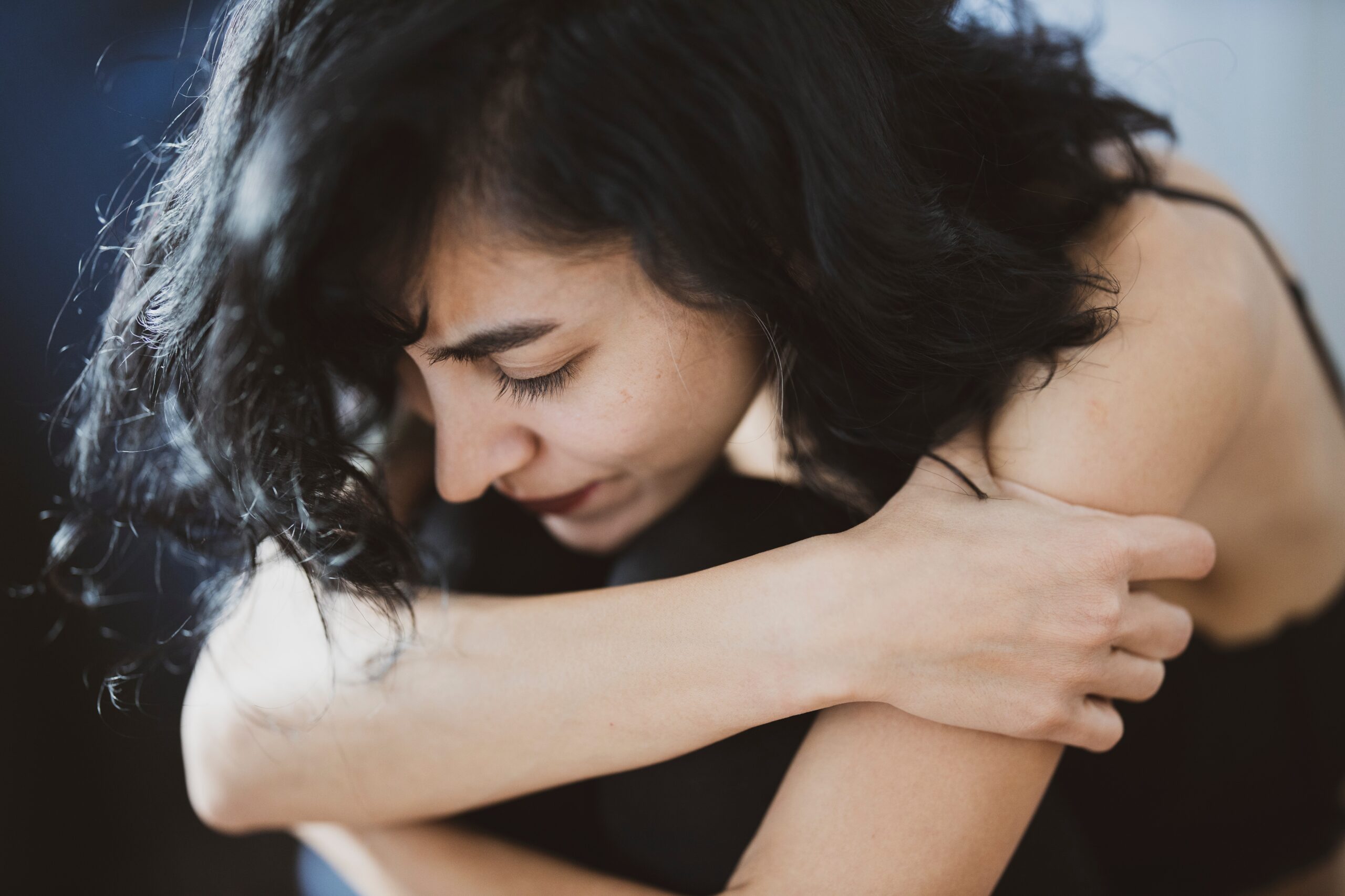 A woman with dark hair sits with her arms wrapped around her knees, appearing thoughtful. Wearing a black top, she closes her eyes, conveying introspection against a softly blurred background. Her pose suggests resilience and strength in the face of overcoming domestic violence.