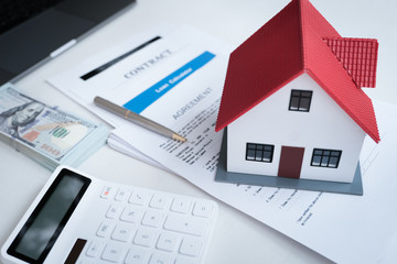 A small model house sits on top of documents labeled "Contract" and "Agreement," next to a stack of cash, a calculator, and a pen, symbolizing real estate, home buying, or issues like mortgage divorce settlements.
