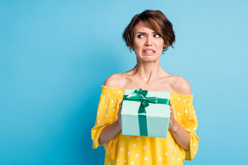 A woman in a yellow polka dot top looks uneasy while holding a wrapped gift with a green ribbon, perhaps reflecting on gifts after divorce, standing against a blue background.