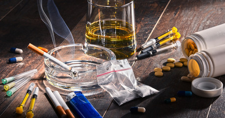 A variety of drugs, including pills, syringes, capsules, a glass of amber liquid, a lighter, a smoking cigarette in an ashtray, and a small plastic bag are scattered on a wooden surface—signs often linked to substance abuse and divorce struggles.