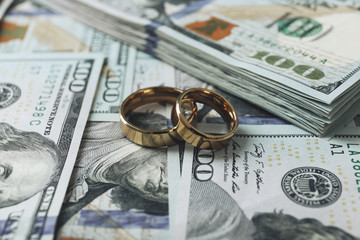 Two gold wedding rings rest on several scattered and stacked U.S. 100 dollar bills, highlighting the connection between marriage and financial matters—and what it truly costs to afford divorce.
