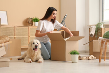 A woman kneels on the floor, unpacking a cardboard box in a bright room. Beside her, under their new pet custody plan, a golden retriever lies contentedly surrounded by moving boxes and plants.