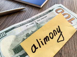 A $100 bill on a wooden table with a yellow sticky note labeled "permanent alimony" in handwritten text, next to a pen and part of a blue notebook.