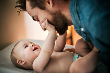 A bearded man in a denim shirt smiles while gently touching noses with a laughing baby, enjoying precious custodial time as the baby reaches up and touches the man's cheek with one hand.
