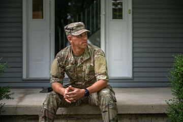 A man in military uniform, possibly reflecting on divorce or a spouse, sits on the front steps of a house with hands clasped. The background features two white doors and gray siding, hinting at personal changes like a military pension coming into play.