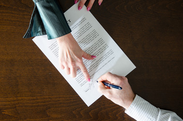 Two people at a table; one with pink nails points to a section on a contract, guiding how to ask a spouse about a postnuptial agreement, while the other holds a pen and is about to sign the document.