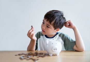A young boy sits at a table with a white piggy bank and scattered coins, examining a coin in his hand with curiosity, perhaps wondering how much child support covers.