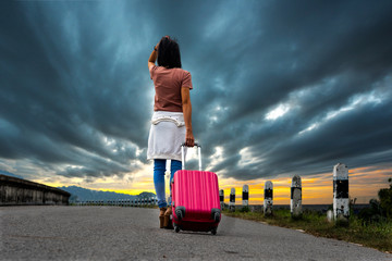 A woman stands on an empty road at sunset, facing away with a pink suitcase. Dark clouds fill the sky, evoking the sense of abandonment and the possibility of a new beginning after divorce.