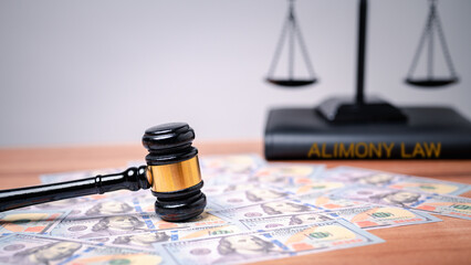 A judge's gavel rests on scattered US hundred-dollar bills, with a law book labeled "Alimony Law" and scales of justice in the background, hinting at questions around whether alimony counts as taxable income.