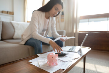 A woman sits on a couch, using a calculator and examining paperwork at a coffee table. A piggy bank, scattered documents, and an open laptop suggest she is learning how to create a budget after divorce.