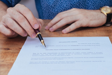 A person wearing a blue shirt and a wristwatch holds a pen over financial affidavits divorce paperwork on a wooden table, appearing ready to sign or review the documents.