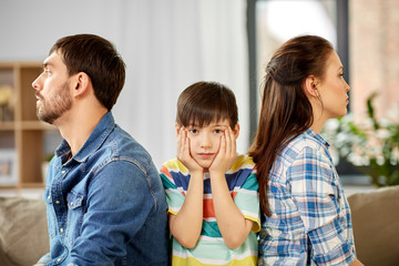 A boy sits between a man and a woman, who are sitting back-to-back with serious expressions, as if making the child choose who to live with. The boy looks worried, his hands on his face.