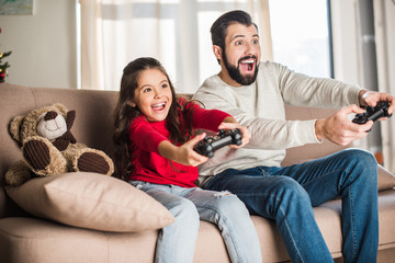 A smiling man and young girl sit on a couch, enthusiastically playing video games with controllers. Despite previously restricted visitation rights, they look excited and engaged, with a brown teddy bear beside them.