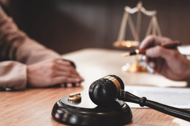 A judge’s gavel, two wedding rings, and legal documents are on a desk. Two people sit across from each other, one holding a pen, with scales of justice blurred in the background.