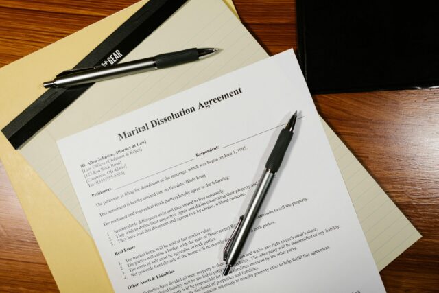 A Marital Dissolution Agreement document on a desk with two pens placed on top, next to a manila folder and a black folder, all set on a wooden surface.