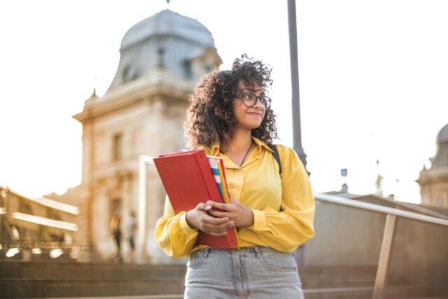 A young woman with curly hair, glasses, and a yellow shirt stands outside her child’s college after divorce in Tennessee, holding red folders and notebooks and smiling. Behind her, a historic building is lit by sunlight.