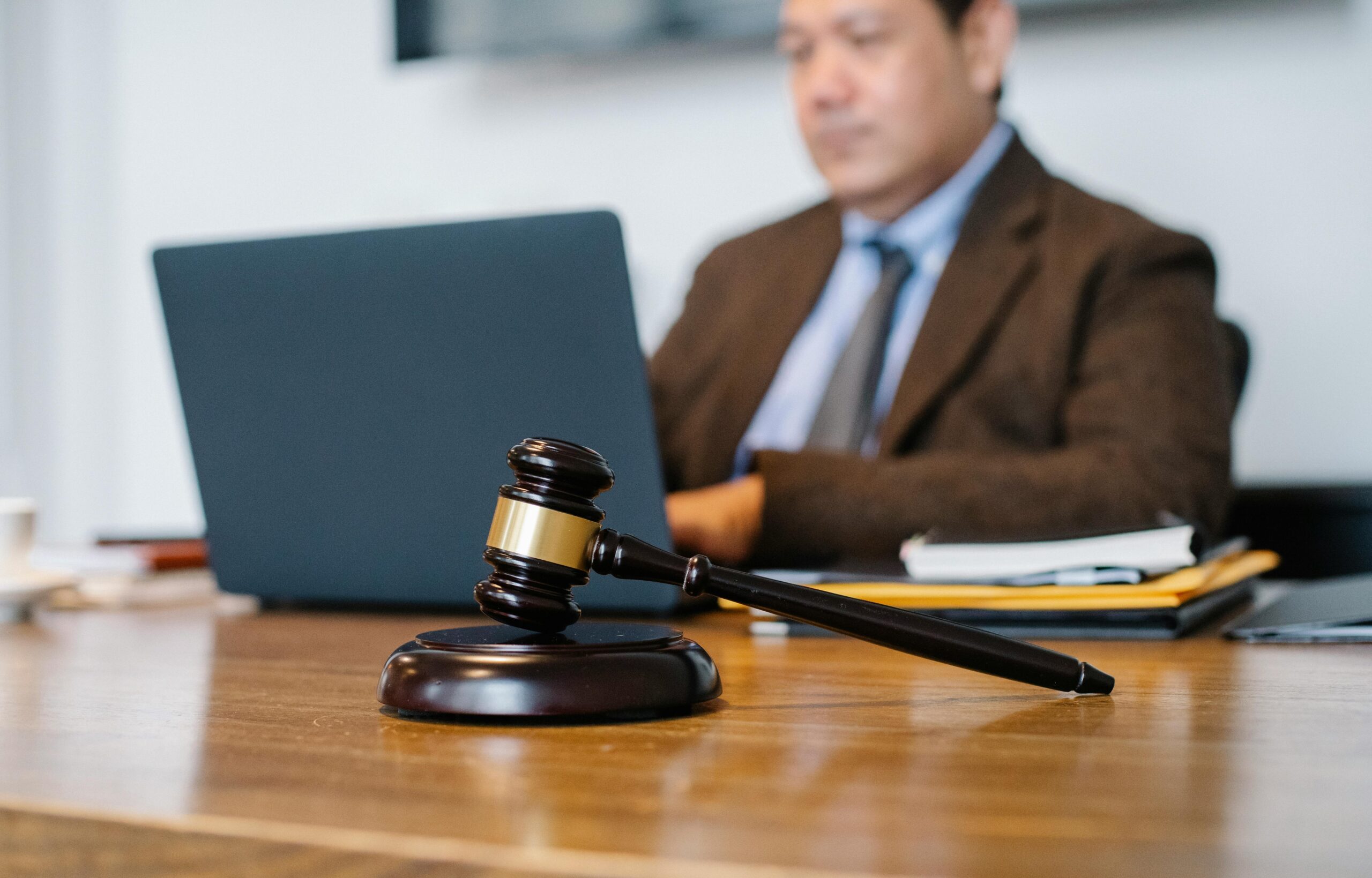 A wooden gavel rests on a desk in the foreground, while a person in a suit works on a laptop in the blurred background.